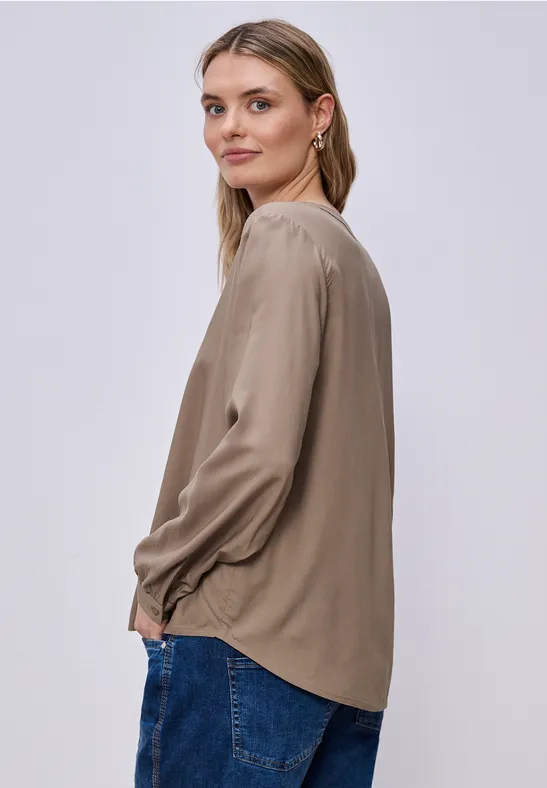 Seitliche Ansicht einer Person in beigem Langarmshirt und blauen Jeans vor hellem, einfarbigem Hintergrund.