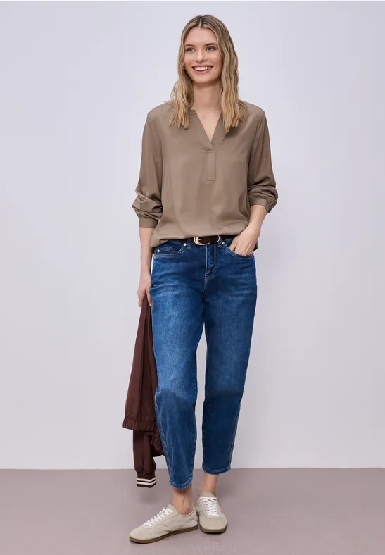 Eine Person in legerer Kleidung: beige Bluse, blaue Jeans, weiße Slip-ons, hält eine braune Jacke.