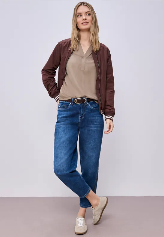 Person in lässigem Outfit aus Jeans, Top, Bomberjacke und Slipper steht entspannt auf hellem Hintergrund.