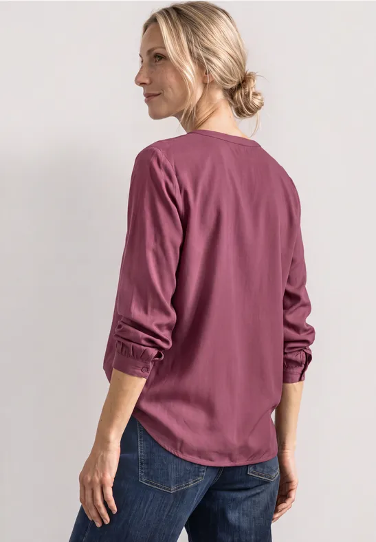 Frau im Profil trägt eine lachsrosa Bluse mit hochgekrempelten Ärmeln und Jeans auf hellem Hintergrund.