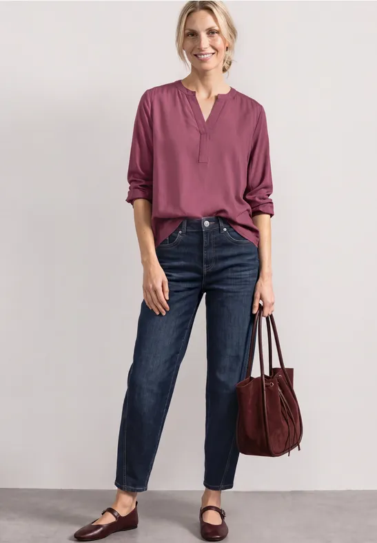 Eine Frau vor neutralem Hintergrund trägt dunkle Jeans, eine plumfarbene Bluse und eine große Handtasche.