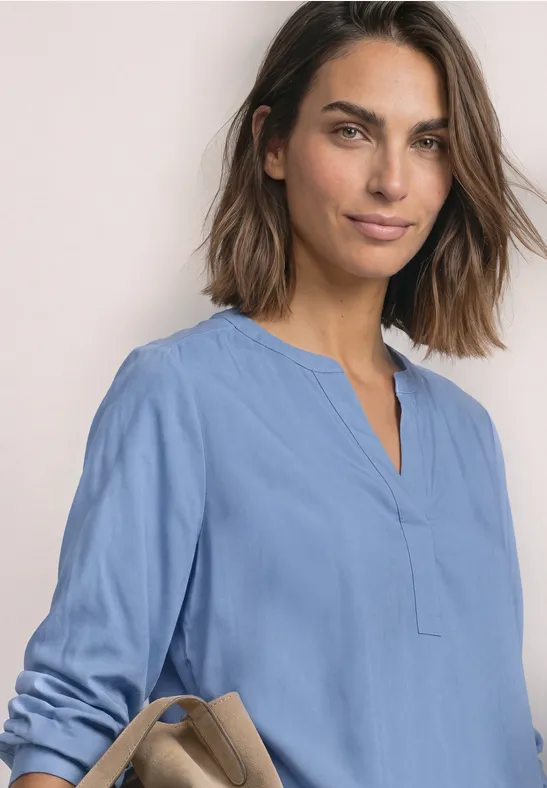 Eine lächelnde Frau mit schulterlangem Haar trägt eine hellblaue Tunika und hält eine beige Handtasche.