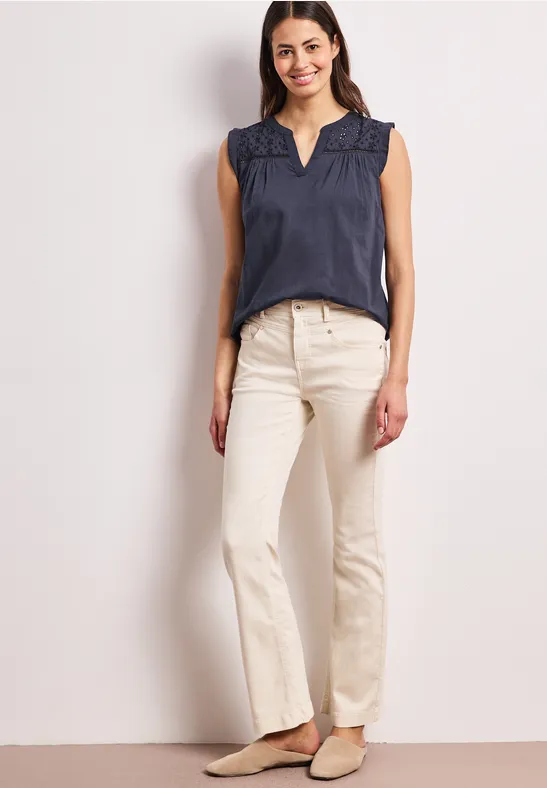Frau trägt ärmellose dunkelblaue Bluse mit Rüschenkragen, weiße gerade Jeans und flache Schuhe.
