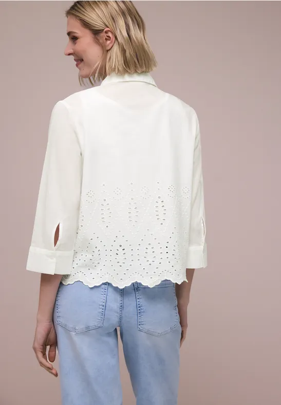 Rugaanzicht van een vrouw in een witte geborduurde blouse met driekwartmouwen en lichtblauwe jeans.