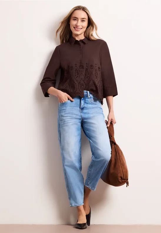 Frau in brauner, bestickter Bluse, hellblauen Jeans, schwarzen Schuhen und brauner Tasche.