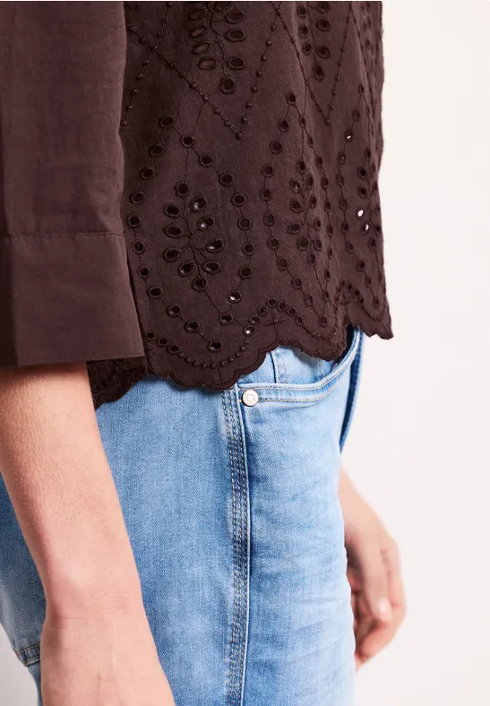 Seitliche Aufnahme einer Person in hellblauen Jeans und brauner Bluse mit Lochstickerei und gewelltem Saum.