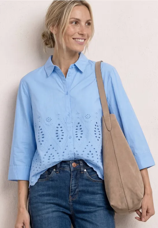 Vrouw in lichtblauwe blouse met open borduursels, spijkerbroek en beige suède tas voor neutrale achtergrond.
