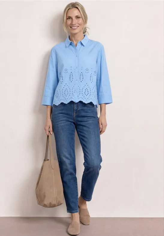 Vrouw in lichtblauw geborduurd overhemd, blauwe jeans, beige mules en taupe suède shopper.