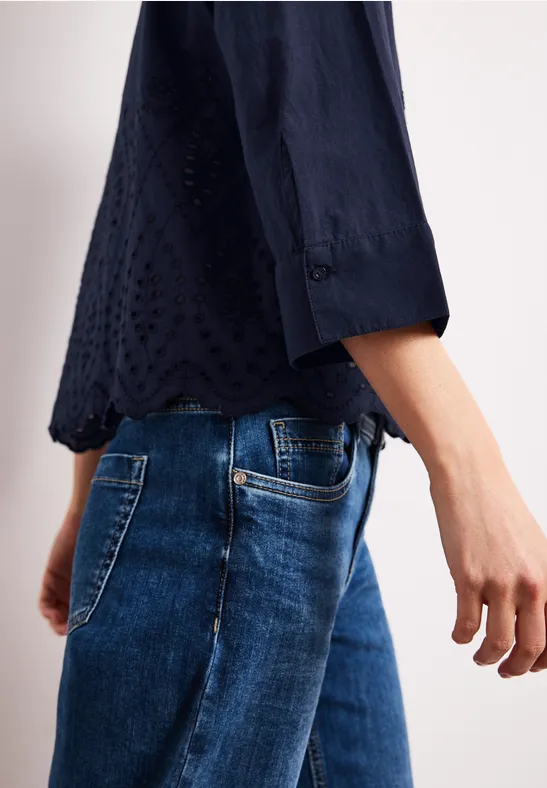 Seitliche Ansicht einer Person in dunkelblauer, bestickter Bluse mit drei Viertelärmeln und blauen Jeans.