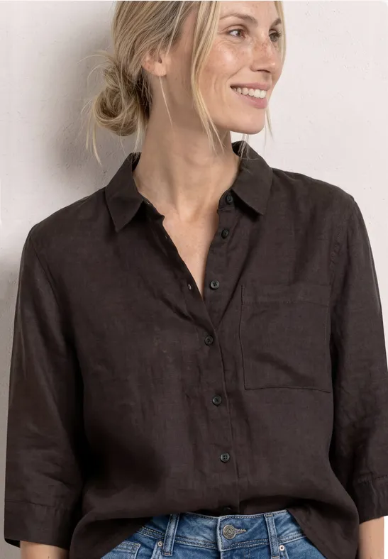 Vrouw in donkerbruine linnen blouse met driekwartmouwen en jeans voor lichte achtergrond.