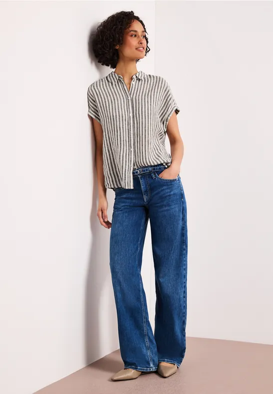 Een persoon leunt tegen een muur, gekleed in een gestreept overhemd en wijde blauwe jeans.
