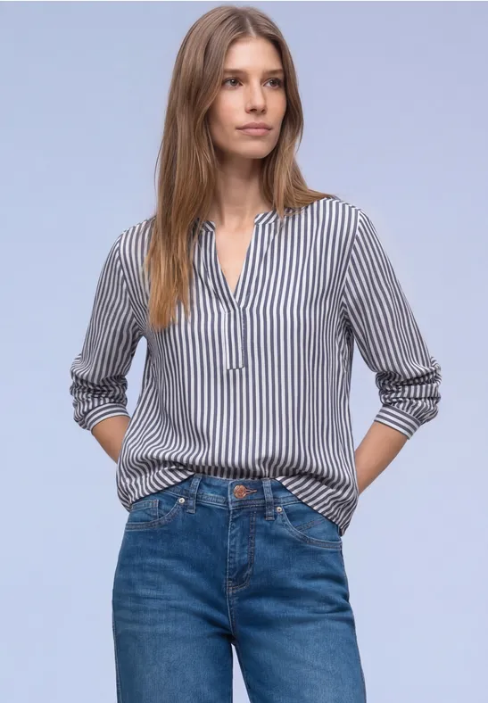 Person steht vor hellem Hintergrund in gestreifter Bluse und blauen Jeans mit Händen in den Hosentaschen.