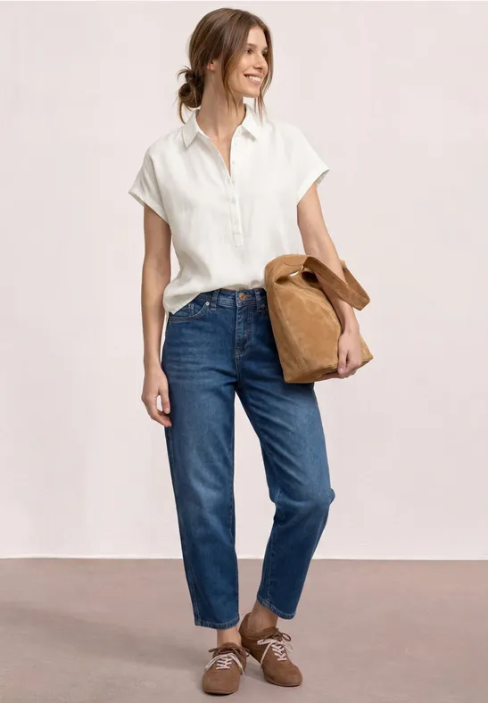 Frau in weißer Bluse, blauen Jeans und braunen Schuhen hält eine braune Tasche.