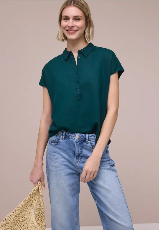 Eine Frau in einer kurzärmligen grünen Bluse und hellblauen Jeans hält eine geflochtene Strohtasche.