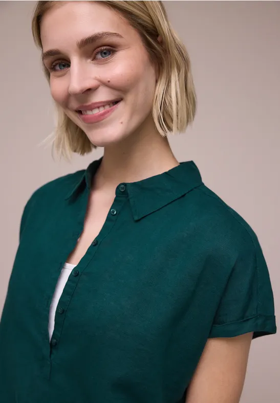 Eine Person mit blonden Haaren in einem dunkelgrünen Kurzarmhemd über einem weißen Top, Gesicht unscharf.