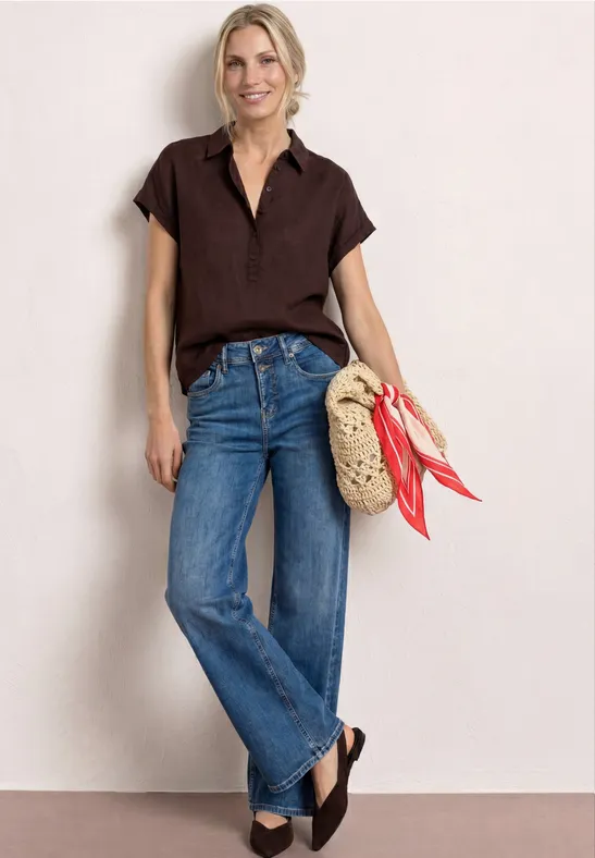 Vrouw in bruin kort shirt en blauwe jeans, met gevlochten rieten tas met rode sjaal en donker sleehakken.