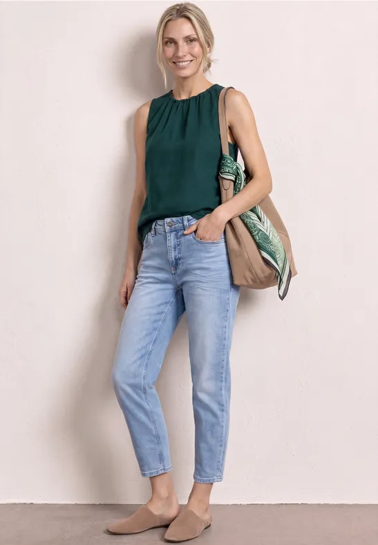 Een vrouw draagt een donkergroen mouwloos topje, lichte jeans, beige slippers en een grote tas.