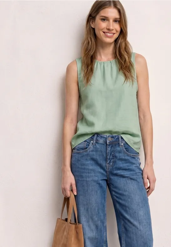 Vrouw in groene mouwloze top en blauwe jeans, met een bruine tas voor een beige muur.