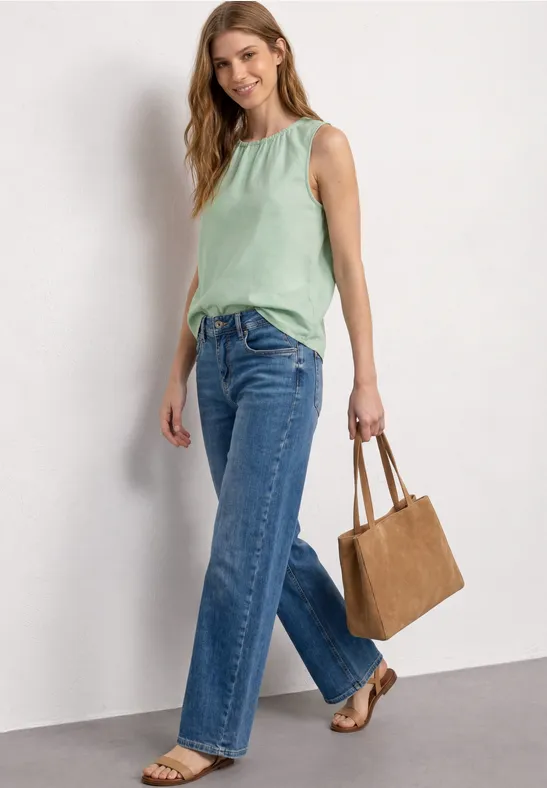 Vrouw in mintgroen top, wijd jeans, bruine sandalen en tas voor een witte muur.