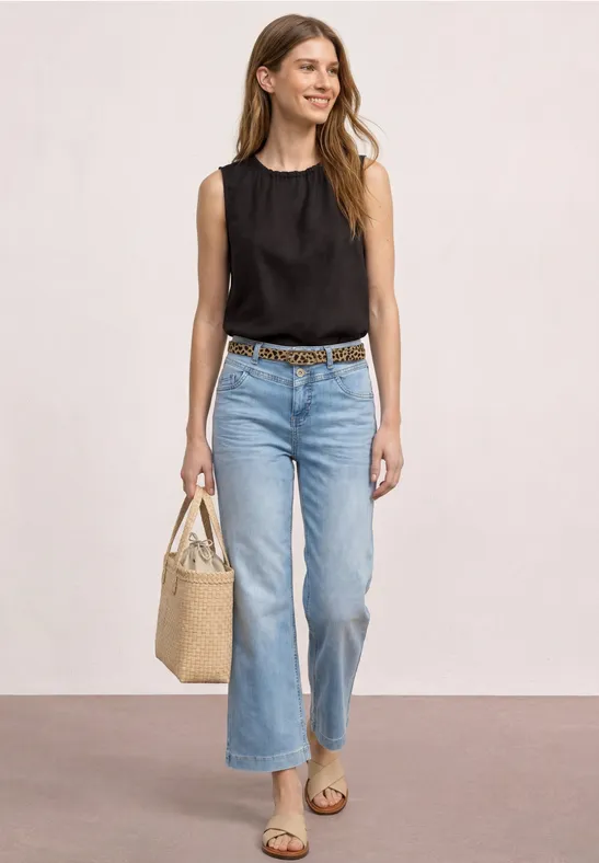 Vrouw met zwarte mouwloze top, lichtblauwe jeans, sandalen en een strooien tas.