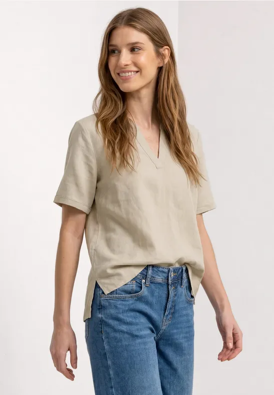Vrouw in beige shirt met korte mouwen en blauwe jeans, staand voor een neutrale achtergrond.