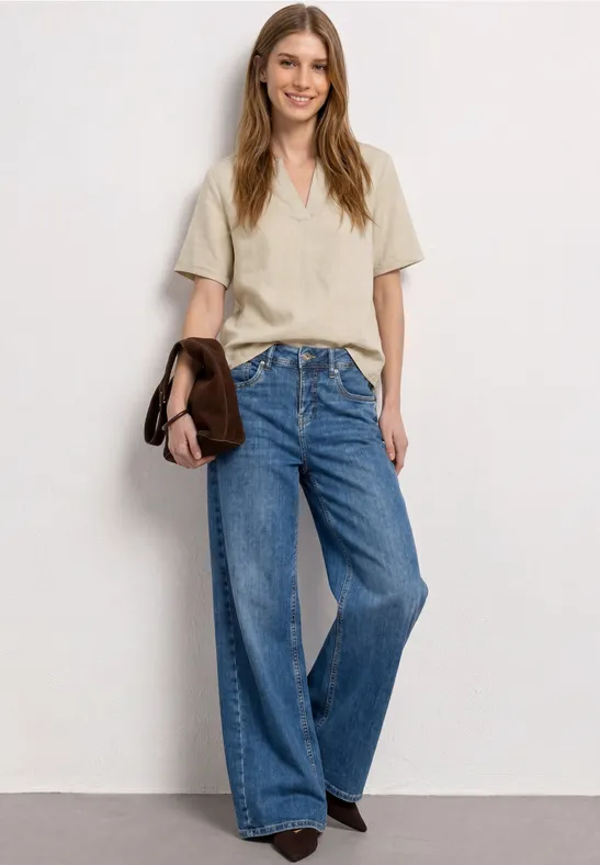 Vrouw in beige T-shirt, wijde jeans en bruine schoenen, met een bruine tas voor een witte achtergrond.