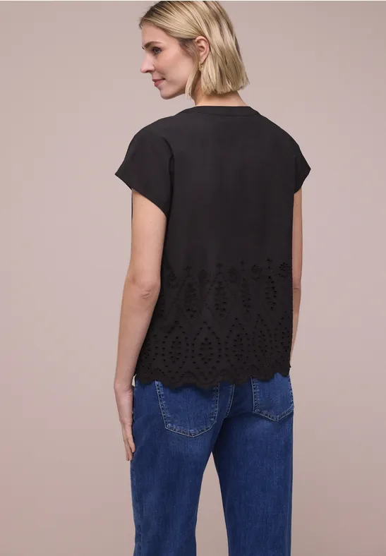 Frau von hinten in schwarzer Kurzarm-Bluse mit floraler Lochspitze am Saum und blauen Jeans vor neutralem Hintergrund.