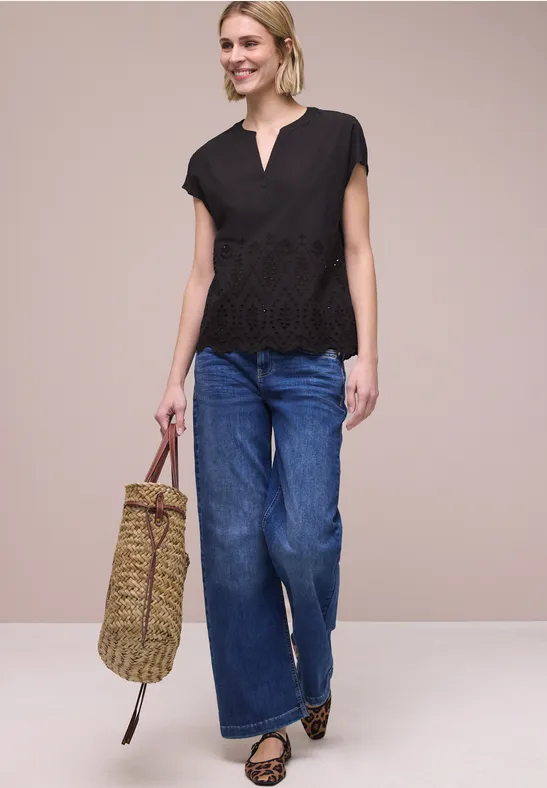 Frau mit schwarzer Spitzenbluse, weiten Jeans, Leoparden-Ballerinas und geflochtener Tasche vor beigem Hintergrund.