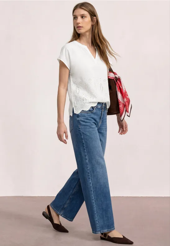 Frau in weißer Kurzarmbluse und weiten blauen Jeans trägt schwarze Flats und hält ein rotes Tuch.