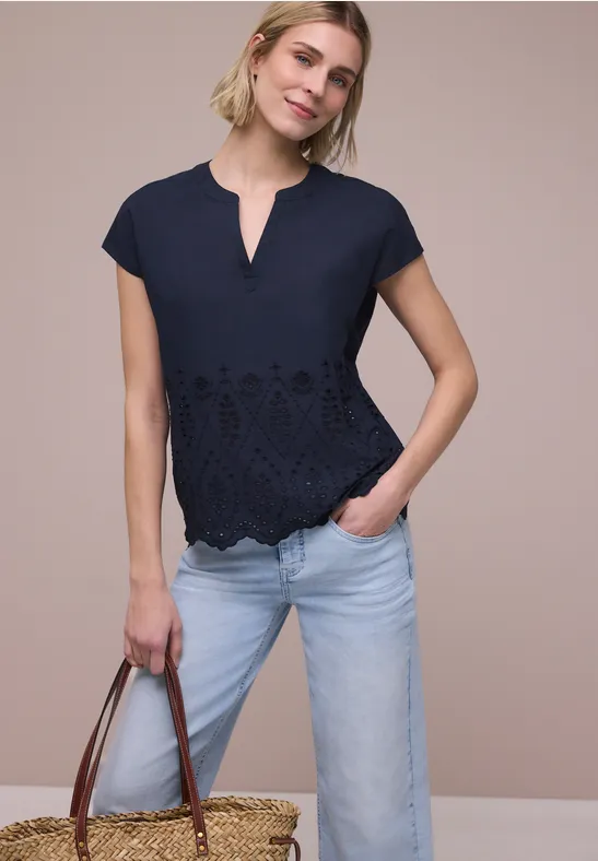 Frau in kurzer, bestickter, dunkler Bluse und heller Jeans hält Ledertasche.