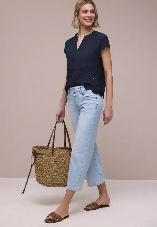Frau in dunkelblauer Bluse, hellblauen Jeans, braunen Sandalen und Korbtasche vor neutralem Hintergrund.