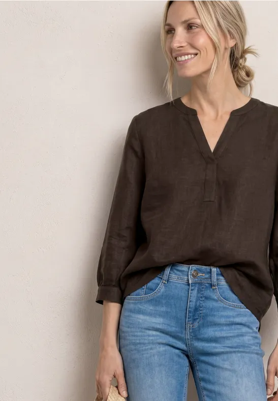 Wazige foto van een vrouw in een bruine linnen blouse met V-hals en blauwe jeans tegen een lichte achtergrond.