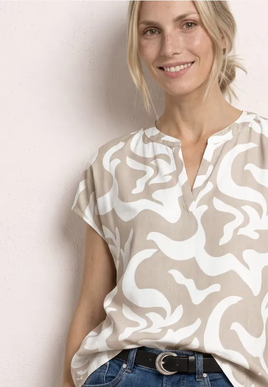 Frau in beige-weiß gemusterter Bluse und Jeans, lächelnd vor neutralem Hintergrund.