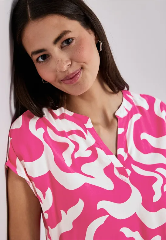 Femme porte un haut rose et blanc à motifs, manches courtes, col en V, cheveux attachés sur un côté.
