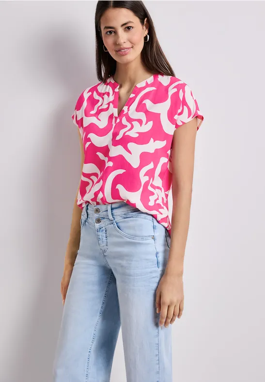Femme en chemisier rose à motifs blancs et jean clair, posant devant un fond neutre.