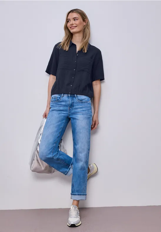 Frau in legerem dunkelblauem Kurzarmhemd, geraden Jeans und Sneakers steht und hält eine Tasche.