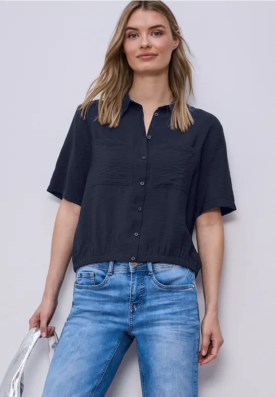 Person trägt schwarze Kurzarmbluse, blaue Jeans und hält eine weiße Handtasche.