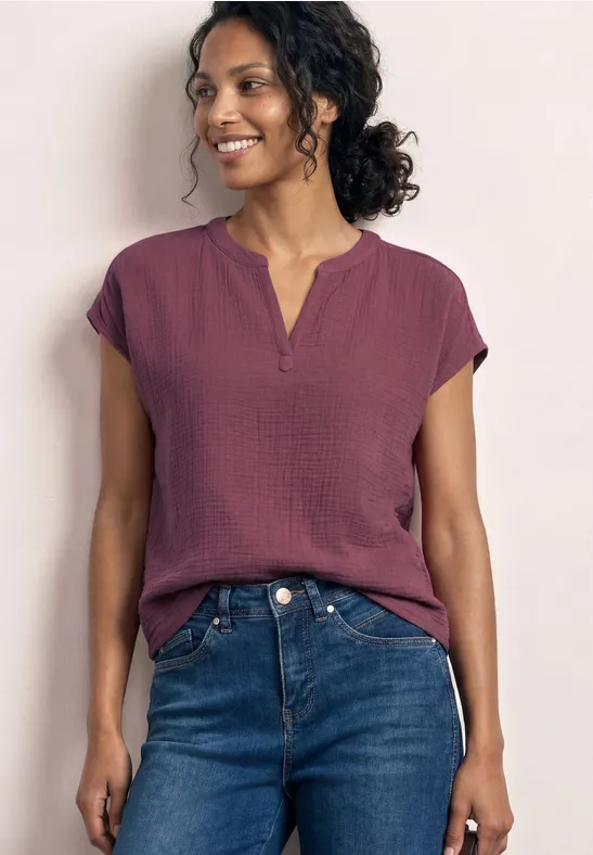 Frau trägt bordeauxrotes Top mit kurzen Ärmeln und blaue Jeans, steht vor hellem Hintergrund.