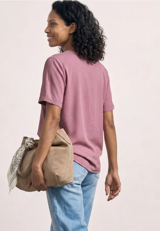 Persoon in pastelroze t-shirt en blauwe jeans, gezien van opzij, draagt een beige tas.