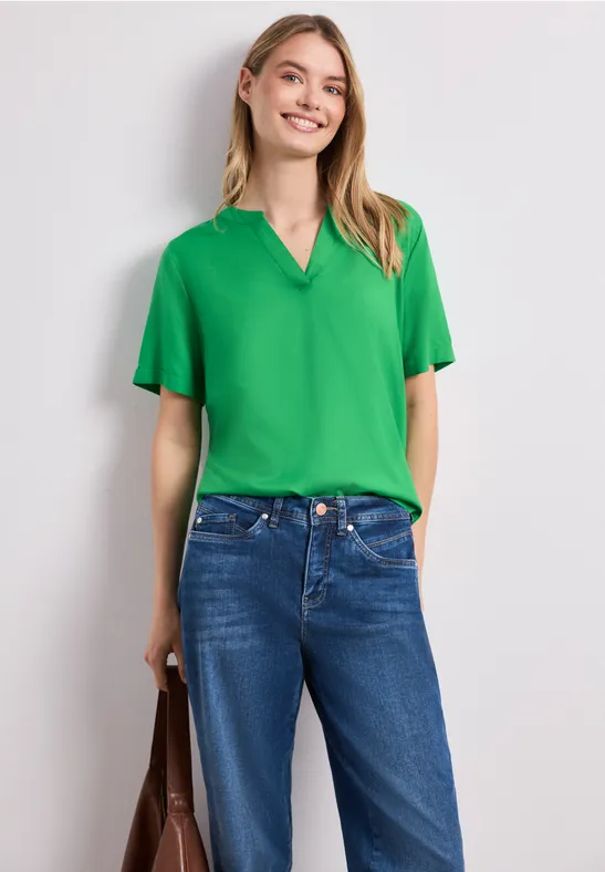 Vrouw in groene blouse, blauwe jeans en bruine tas voor een neutrale achtergrond.