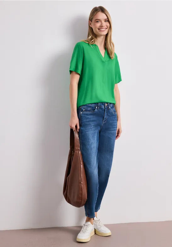 Vrouw draagt groen T-shirt, blauwe jeans, witte sneakers en grote bruine tas voor een lichte achtergrond.