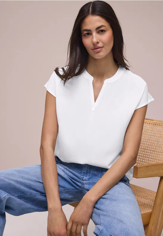 Een vrouw in een wit shirt en blauwe jeans zit op een stoel met gevlochten rug, handen in de schoot.