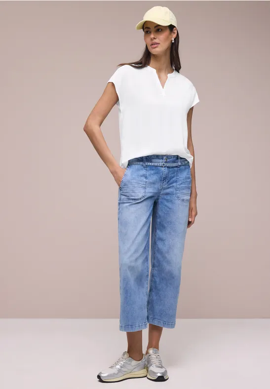 Vrouw in wit shirt, wijde blauwe jeans en zilveren schoenen voor een neutrale achtergrond.