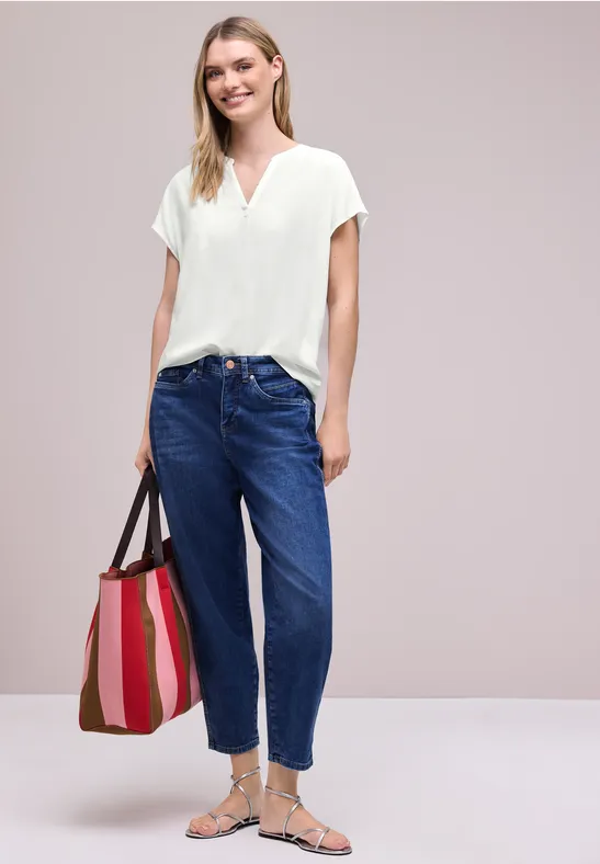 Femme en chemisier blanc à col V, jean taille haute bleu, sandales argentées et sac cabas rayé.