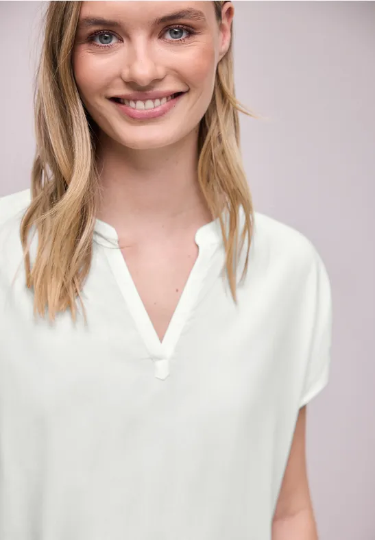 Une personne porte un chemisier blanc à col en V sur un fond neutre.