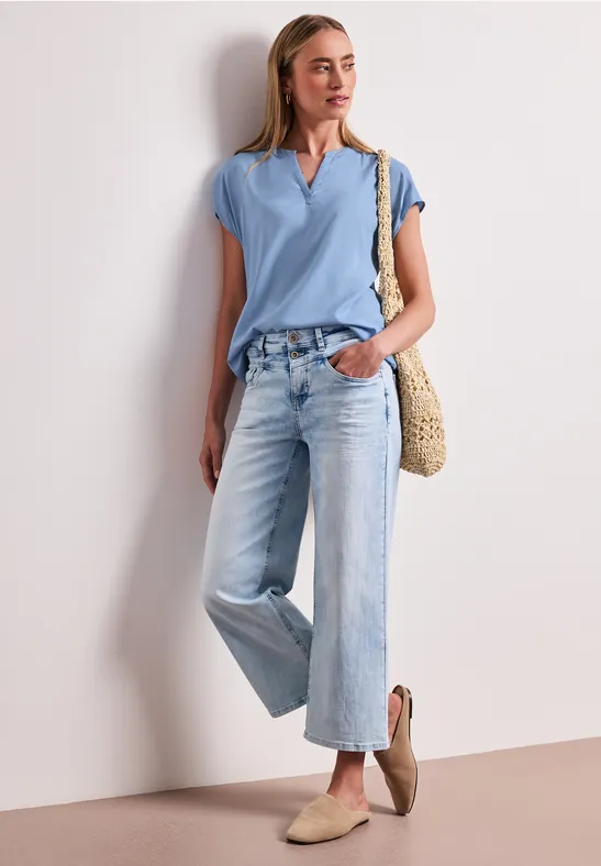 Frau in hellem T-Shirt, weitem Jeans, Basttasche und beigen Slippern lehnt an weißer Wand.