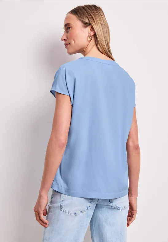 Eine Frau von hinten in hellblauem T-Shirt und Jeans, Haare locker, lässige Haltung.