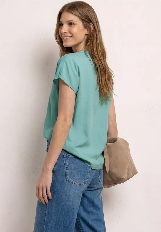 Frau mit türkisfarbenem T-Shirt, blauer Jeans und beiger Tasche, Blick über die Schulter.