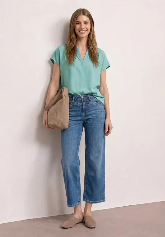 Frau vor heller Wand, trägt mintgrünes T-Shirt, weite blaue 7/8-Jeans, beigen Shopper und Loafer.