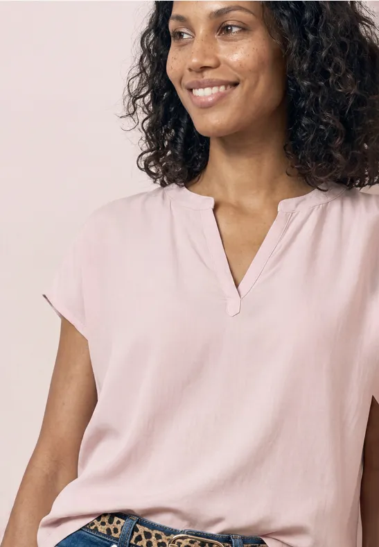 Bovenlichaam van een persoon met krullend haar, roze blouse met V-hals, lichte achtergrond.